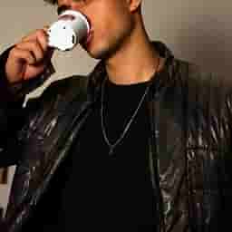 Man wearing a black leather jacket and necklace sipping from a small white cup.