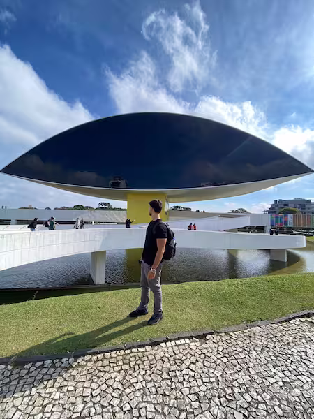 Person standing on a grassy area in front of the Museu Oscar Niemeyer with its distinctive large eye-shaped structure and white bridge over water.