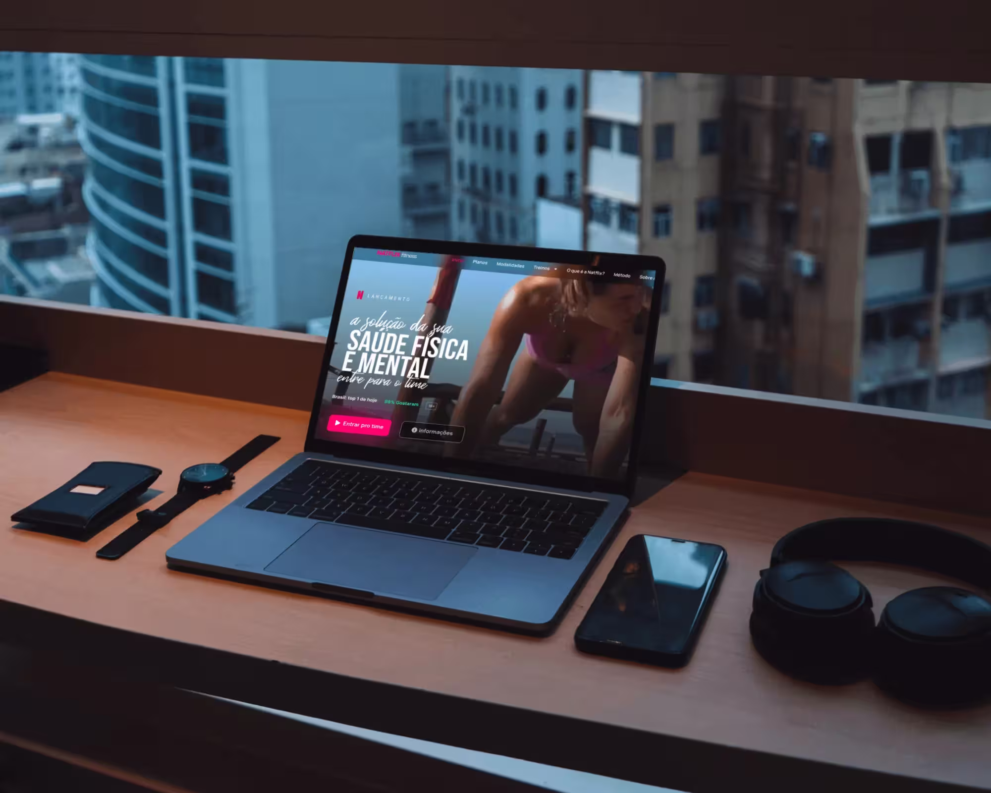 Laptop on a wooden desk showing a fitness website with a woman exercising, beside a smartphone, smartwatch, wallet, and headphones near a window overlooking city buildings.